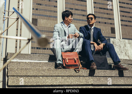Two business partners sitting on the steps outdoors and talking. Korean business men discussing work while sitting on stairs outside in the city. Stock Photo