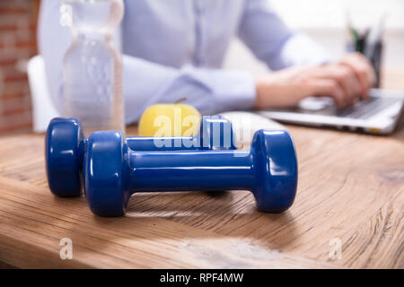Businessperson Working On Laptop Sitting Near Water Bottle And Blue Dumbbells On Office Desk Stock Photo