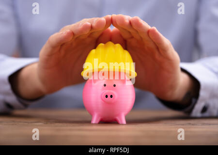 Close-up Of A Person's Hand Protecting Pink Piggy Bank With Yellow Hard Hat Stock Photo