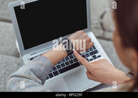 Time management. Young lady sitting on the city street checking time on watch back view close-up Stock Photo