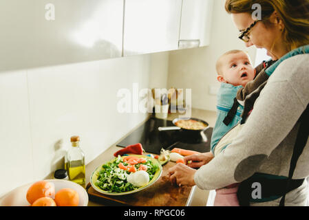 Smiling baby ported in baby carrier backpack looking at his mother while she cooks, concept of family conciliation Stock Photo