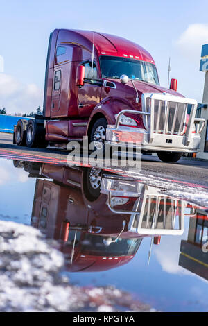 Red big rig semi truck tractor for industrial freight in American logistic system with aluminum bumper grille protector reflecting in puddle of melted