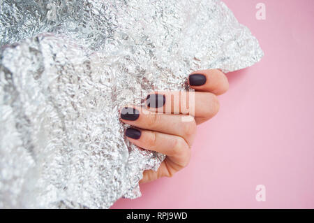 Young woman's hand with beautiful manicure on pink background holding metallic shiny paper. Stock Photo