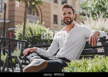 Portrait of handsome young brown-haired man sitting on a bench in the park and looking away. Man dressed in a white shirt and jeans with a bicycle beside him. Summertime, relax Stock Photo