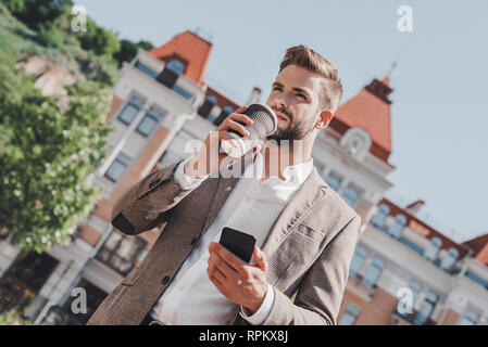 Young bearded businessman stands on city street, holds cup of coffee and uses smartphone. There is building on background. Man working, blogging, chatting online. Social media. Stock Photo