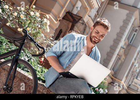 Portrait of handsome young man sitting in the park and holding his laptop. Bearded brown-haired man dressed in a blue striped shirt and jeans looking in his laptop and smiling. Summertime, relax Stock Photo