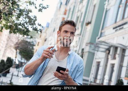 Young stylish man wearing blue shirt and grey T-shirt is walking outdoors, listening to music and looking aside. He holds his phone while moving fast Stock Photo
