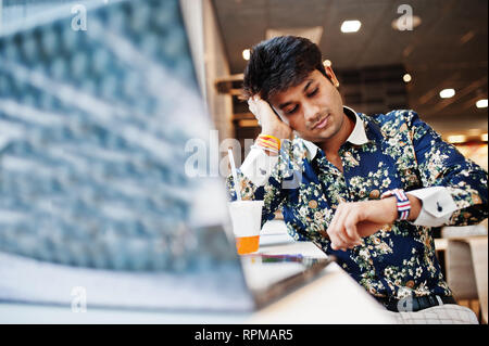 Young male indian freelancer sitting open laptop computer in fast food cafe, handsome asian man looking at watches during work on net-book in comforta Stock Photo