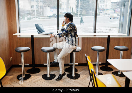 Young male indian freelancer sitting open laptop computer in fast food cafe, handsome asian man speaking phone during work on net-book in comfortable  Stock Photo