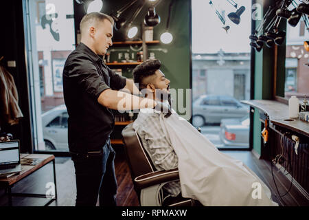 Handsome bearded man is smiling while having his hair cut by hairdresser at the barbershop Stock Photo