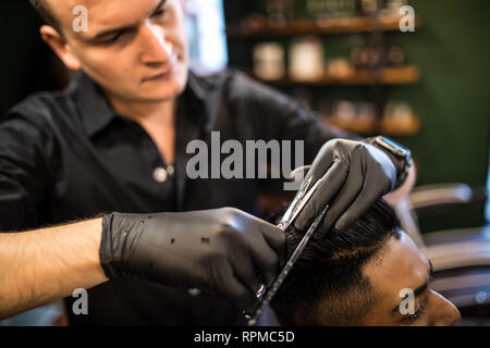 Close up of man getting trendy haircut at barber shop. Male hairstylist serving client, making haircut using machine and comb.
