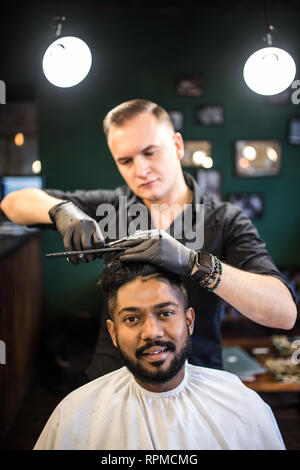 Hairdresser cuts hair, with scissors and black comb, on crown of handsome satisfied client on barber shop Stock Photo