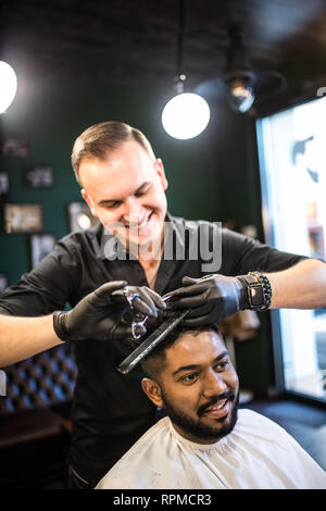 Hairdresser cuts hair, with scissors and black comb, on crown of handsome satisfied client on barber shop Stock Photo