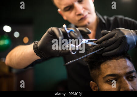 Close up of man getting trendy haircut at barber shop. Male hairstylist serving client, making haircut using machine and comb.