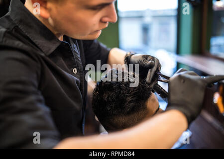 Close up of man getting trendy haircut at barber shop. Male hairstylist serving client, making haircut using machine and comb.