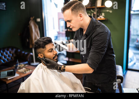 Professional styling. Close up side view of young bearded man getting haircut by hairdresser with electric razor at barbershop Stock Photo