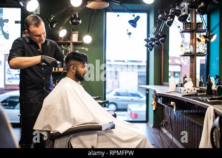 Unshaven man being clipped with professional electric shearer machine in barbershop. Male beauty treatment concept. Indian guy trim beard and mustache Stock Photo