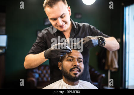 Unshaven man being clipped with professional electric shearer machine in barbershop. Male beauty treatment concept. Indian guy trim beard and mustache Stock Photo