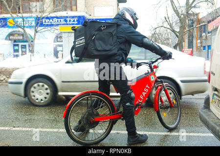 Pizza courier in helmet on the red electric bike tries to squeeze in between cars on the winter ice covered slippy road on his way to clients in Kiev Stock Photo