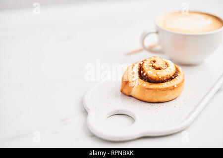 Freshly baked cinnamon roll with spices and cocoa filling and coffee or cappuccino with latte art on white serving plate over white marble background. Stock Photo