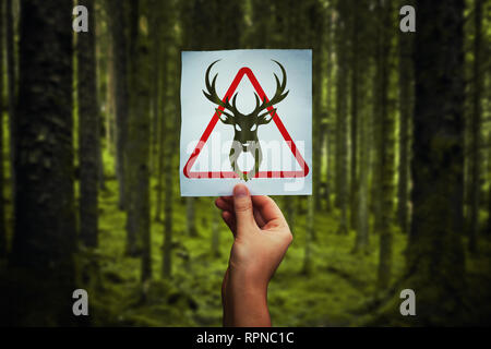 Animal poaching restricted as hand holding a paper sheet with deer symbol over forest background. Red restriction triangle, stop hunting. IUCN list of Stock Photo