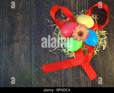Four painted Easter eggs in a nest of straw with a flower in the center, lying on a scarlet ribbon on a wooden table. Close-up. Stock Photo