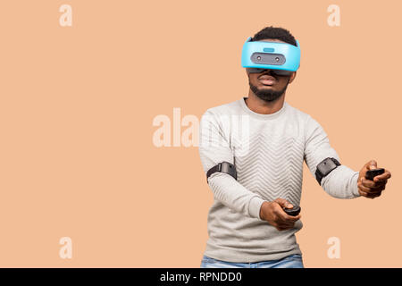 Studio shot of African american man using virtual reality headset, operating with joystick in hands, moving over beige background Stock Photo