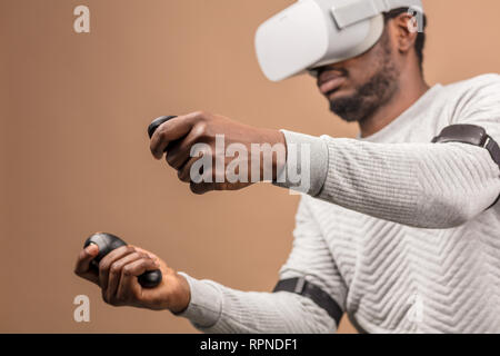Dark-skinned millennial tourist in casual clothing wearing glasses with head-mounted display for smart phone, operating with controllers in hands, doi Stock Photo