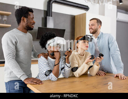 Multicultural group of friends playing video games wearing virtual reality glasses with controllers, women trying to play and boyfriends making hints Stock Photo