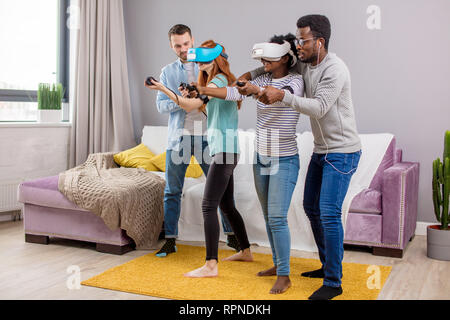 Multicultural group of friends playing video games wearing virtual reality glasses with controllers, women trying to play and boyfriends making hints Stock Photo