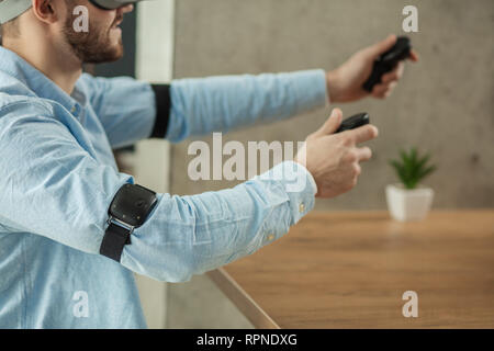 Side view of bearded caucasian man in blue shirt wearing VR glasses and holding controllers in hands while sitting at the table in room. Stock Photo