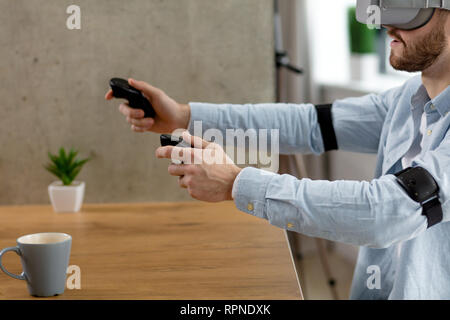 Side view of bearded caucasian man in blue shirt wearing VR glasses and holding controllers in hands while sitting at the table in room. Stock Photo