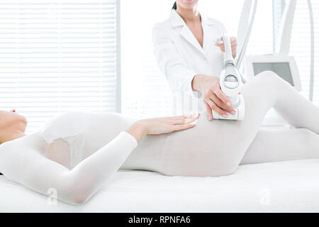 Unrecognizable female beautician in white medicine gown, attends female patient in cosmetological wellness clinic, performing LPG massage on hips in w Stock Photo