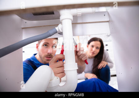 Young Woman Looking At Male Plumber Fixing Sink Pipe In Kitchen Stock Photo