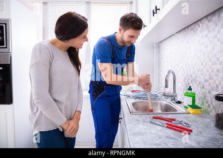 Happy Woman Looking At Male Plumber Using Plunger In The Kitchen Sink Stock Photo
