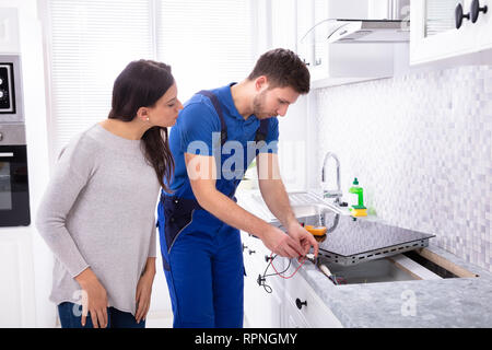 Male Repairman Installing Induction Cooker With Woman Standing In Kitchen Stock Photo