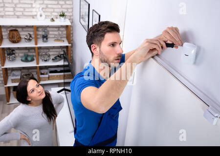 Happy Woman Looking At Electrician Installing Security System Motion Detector On Wall Stock Photo