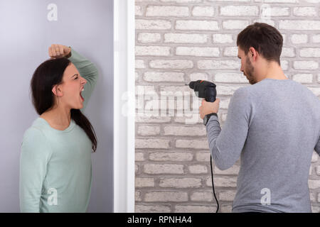 Woman Shouting Behind The Wall With Neighbor Man Using Power Drill On White Brick Wall Stock Photo