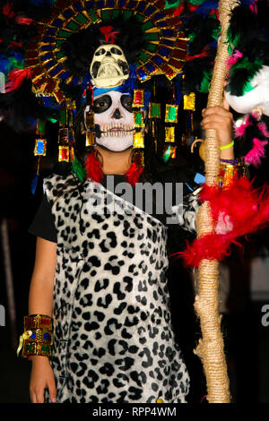 Young boy heavily disguised, with colorful feathers and maya skull mask, wearing leopard outfit, standing in the street during the annual mexican cele Stock Photo