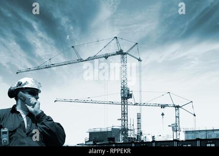 Construction worker talking on mobile phone, with cranes and scaffolding in the background. Stock Photo