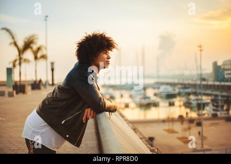 Young man with afro hairstyle standing at a handrail looking sunset with happy relaxed expression Stock Photo