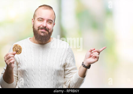 Young hipster man eating chocolate chips cookie over isolated background very happy pointing with hand and finger to the side Stock Photo