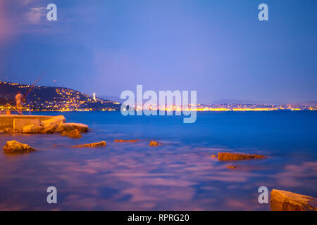 Vittoria Light, Trieste by night. Blue and purple dusk, city lights, Adriatic Sea coastline, rocks, distant lighthouse. Vibrant colors. Long exposure Stock Photo