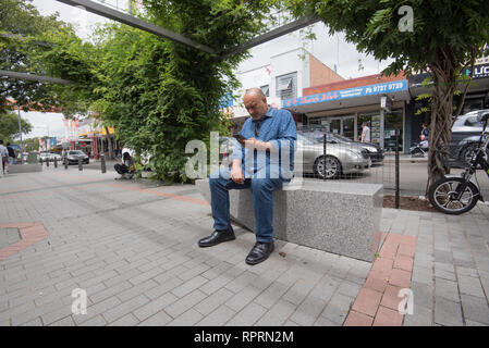 A balding middle-aged, sub-continent man in his late 40's sits concentrating while reading from his phone in John Street Cabramatta in Sydney Stock Photo