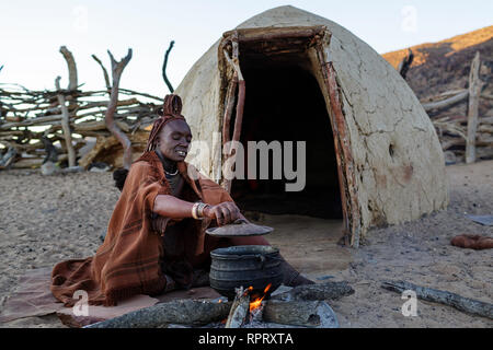 Himba woman cooking dinner on fire outside the mud hut. Purros, Nambia, Africa Stock Photo