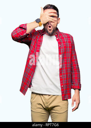 Handsome man wearing casual shirt peeking in shock covering face and eyes with hand, looking through fingers with embarrassed expression. Stock Photo
