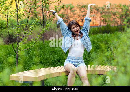 Portrait of beautiful young brunette woman in blue casual denim style sitting and outdoor spring, yawning with closed eyes and raised arms. outdoor sp Stock Photo