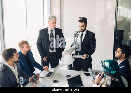 Male caucasian employee presents business strategy on white paper shift to top managers of his company in boardroom. Chief is standing beside, all the Stock Photo