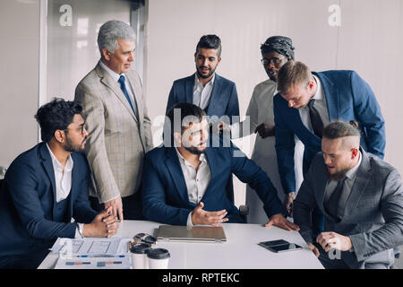 Confident team of age and race diverse male professionals dressed in formal wear excited with success of construction company gatther around boss tabl Stock Photo