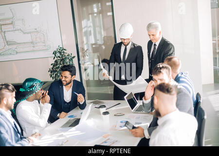 Multiracial group of constructors, builders, engeneers and architects discussing blueprint at office. All men are dressed in business suits, and white Stock Photo